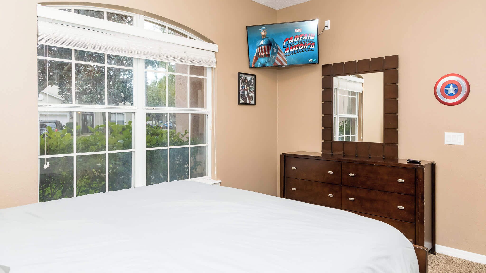 A bedroom with a large bed, a dresser, a TV mounted above the dresser displaying 'Captain America', a window with blinds, and a Captain America shield on the wall.