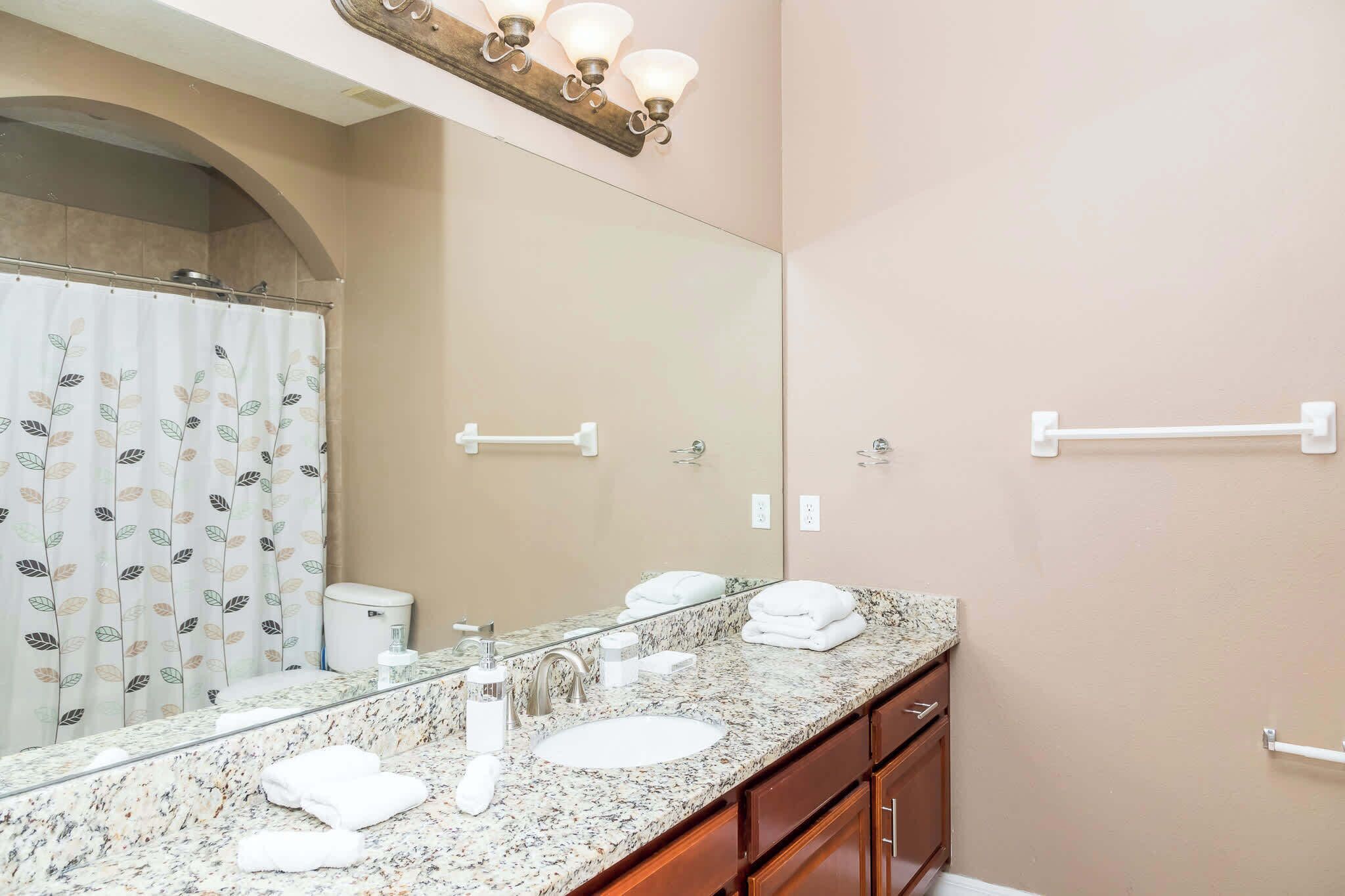 Une salle de bain dotée d’un grand miroir, d’un comptoir en granit, d’un lavabo blanc, d’un rideau de douche orné de motifs de feuilles et d’un porte-serviettes.