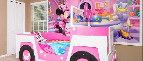 A pink and white playroom with Minnie Mouse and Daisy Duck decorations, a window with blinds, and a white door.