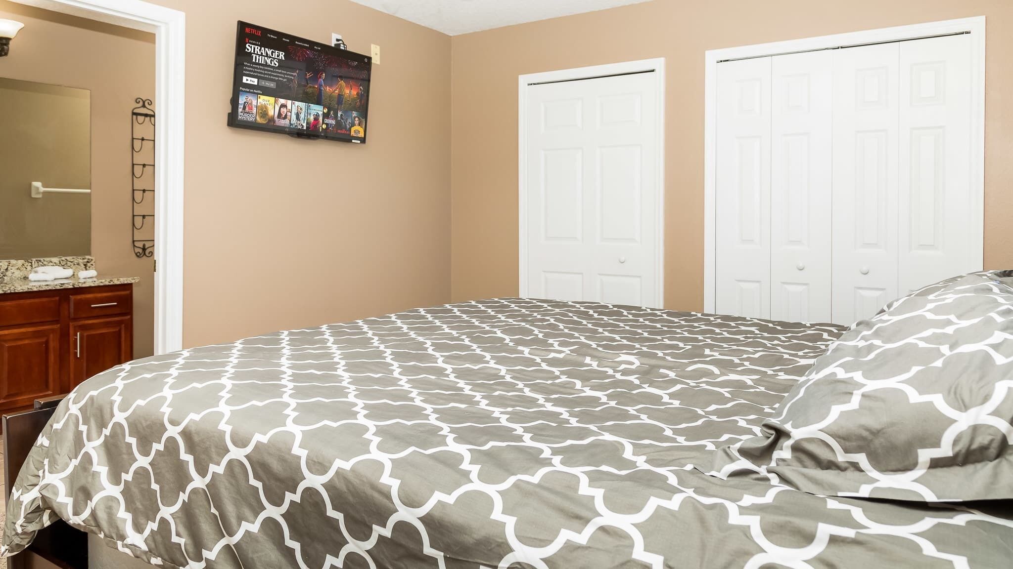 A bedroom with a bed, a dresser, a mirror, and a wall-mounted TV displaying a Netflix show.