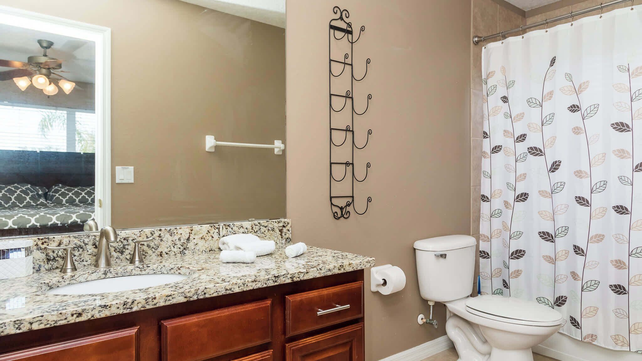 Une salle de bain avec un comptoir en granit, des toilettes, un rideau de douche orné d’un motif de feuilles et un porte-serviettes fixé au mur.