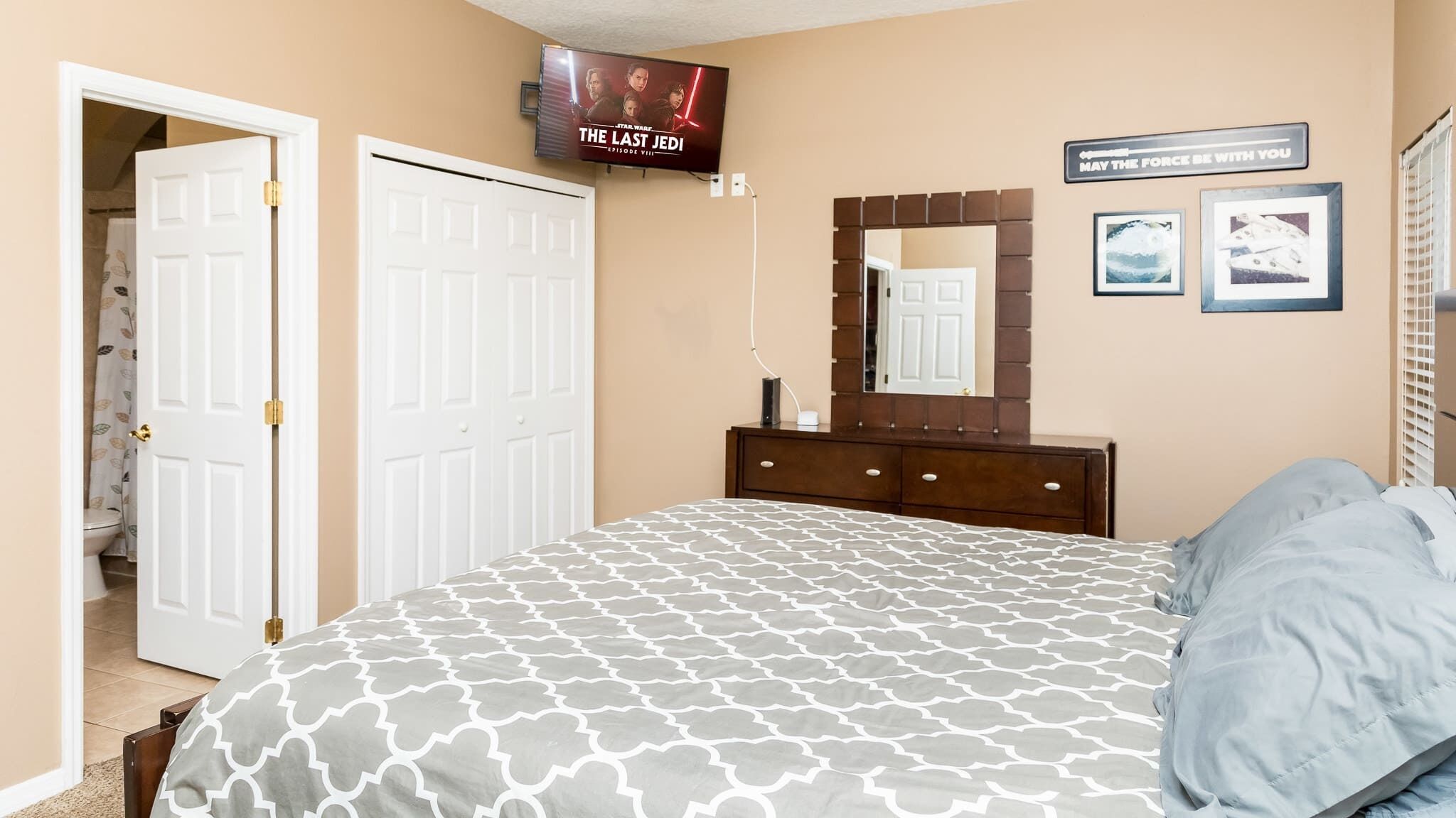 Un dormitorio con una cama, televisor montado en la pared, cómoda y puerta que da a otra habitación.