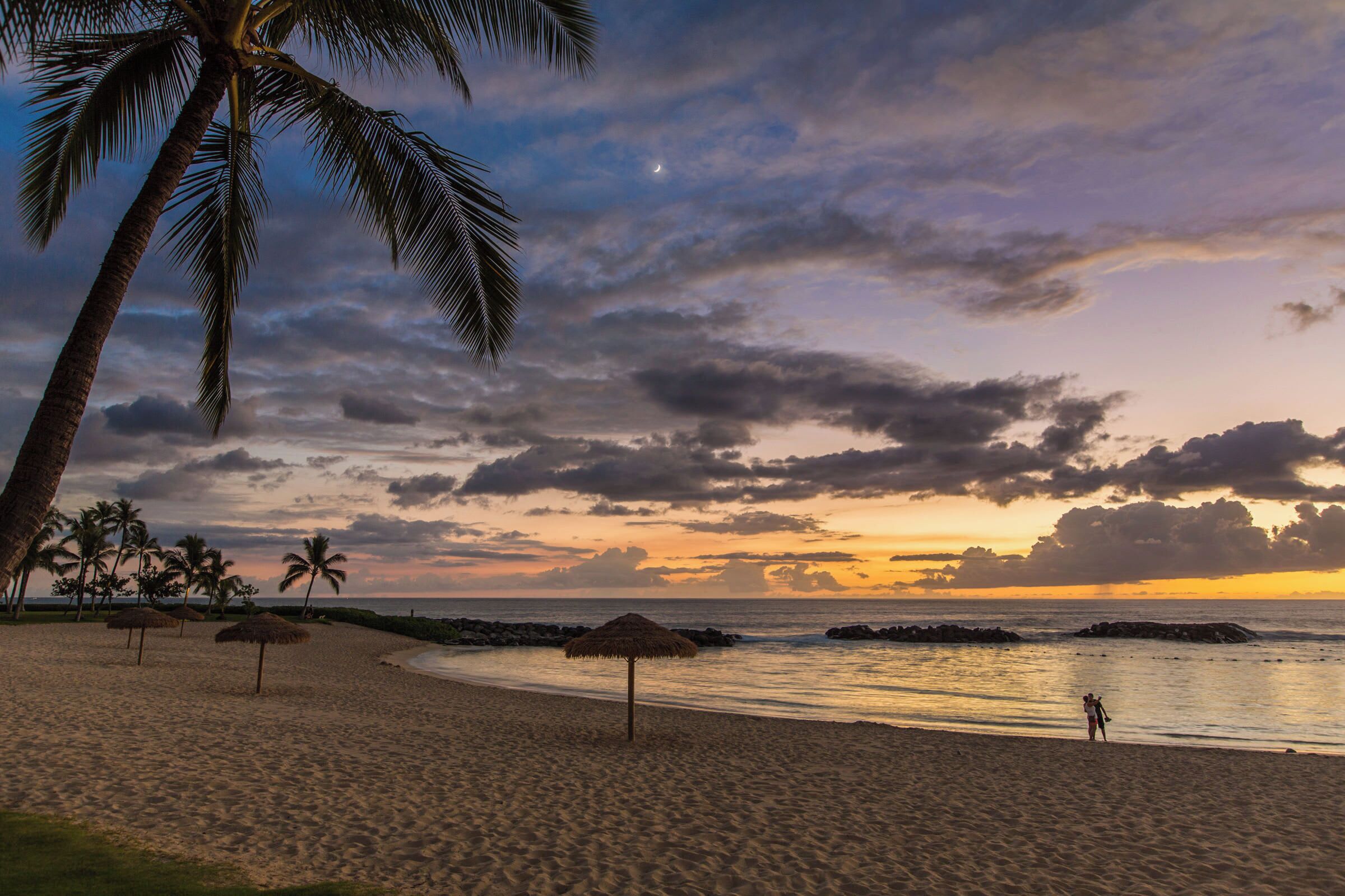 Ein Strand mit Palmen, einem Reetdachschirm und einem ruhigen Meer bei Sonnenuntergang.