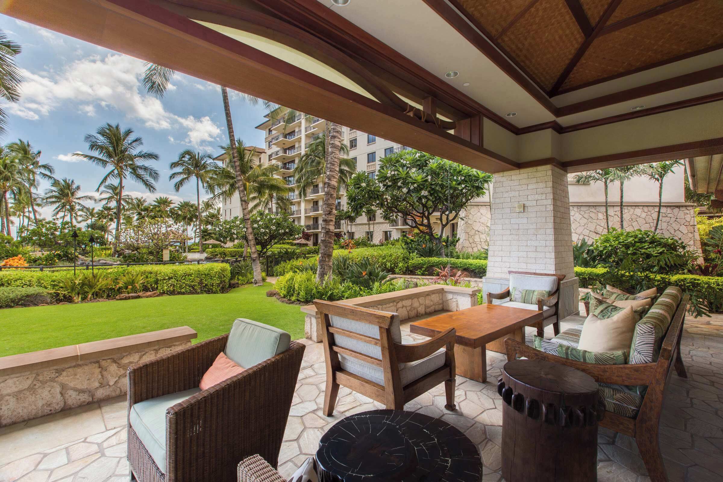 An outdoor patio area with wicker furniture, a stone table, and a view of a well-maintained garden and buildings in the background.