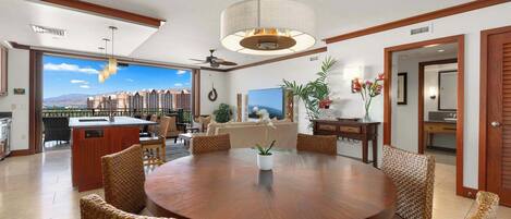 Uma sala de estar ampla com uma mesa de madeira redonda, cadeiras de vime, um ventilador de teto e uma grande janela com vista para a paisagem urbana.