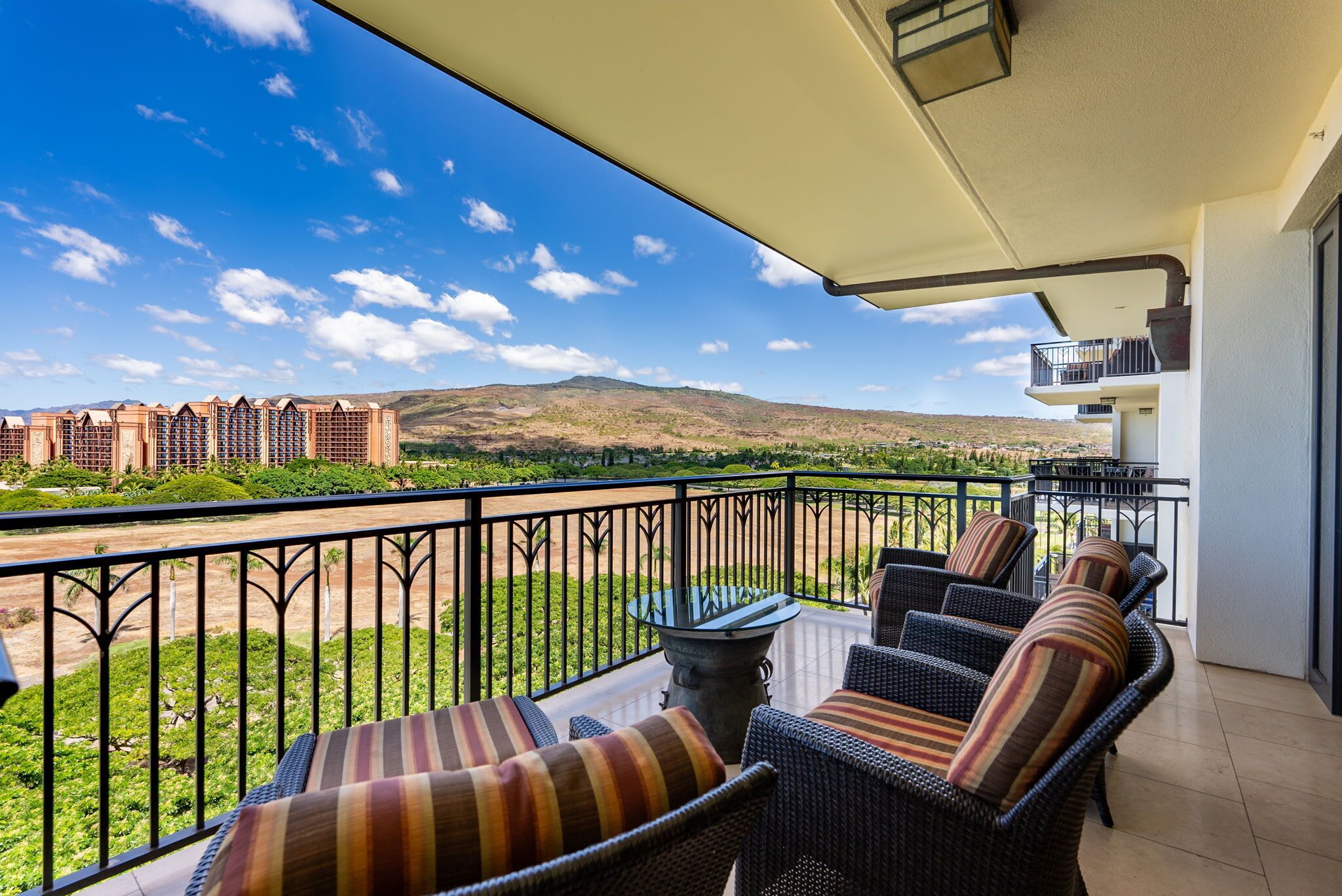 A balcony with wicker furniture and a glass table, overlooking a landscaped area and distant buildings.