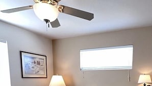 Een slaapkamer met een bed, een plafondventilator, een raam en een wandlamp.