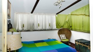 A bedroom with a bed, a chair, a lamp, a window with curtains, and a green suitcase. - Romantic attic with breathtaking views in the old town centre. (Napoli)