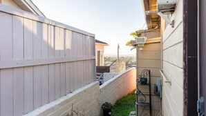 Une ruelle étroite, bordée d’une clôture en bois d’un côté et d’un mur de briques de l’autre. On y trouve un petit espace jardin avec une plante en pot et une étagère en métal sur laquelle sont disposés divers objets.