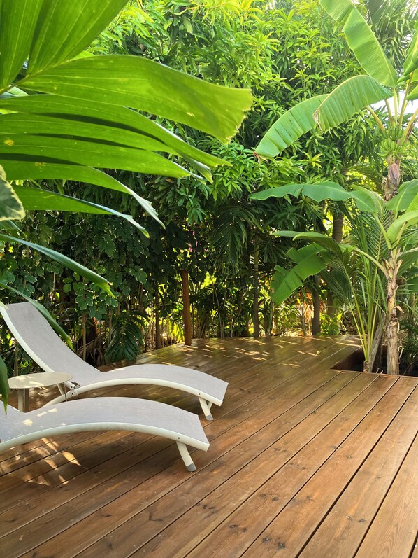 A wooden deck with two white lounge chairs and a small table, surrounded by lush green foliage.