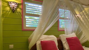 A room with a bed, a bedside table, a wall-mounted light fixture, and a window with a view of greenery. - Zikak - Charming Creole cottage 2 steps from the sea (Sainte-Anne)