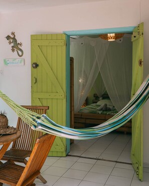 A room with a green door, a hammock, a bed with a mosquito net, and a small table with a pineapple and woven items.