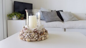 Een witte tafel met een glazen kandelaar met een kaars en schelpen, een witte bank met grijze kussens, en een kleine potplant met gele bloemen.