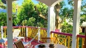 A covered outdoor seating area with a table and chairs, surrounded by a railing with yellow and pink accents, and a view of greenery and trees. - Gwozey - Charming Creole studio 2 steps from the sea (Sainte-Anne)