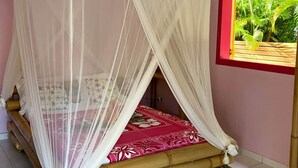A room with a bed under a white canopy, a red-framed window with a view of greenery, and a tiled floor. - Gwozey - Charming Creole studio 2 steps from the sea (Sainte-Anne)