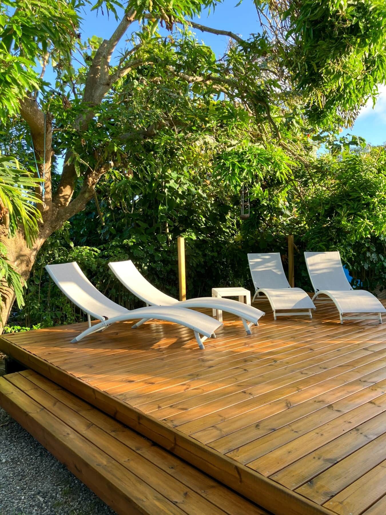 A wooden deck with four white lounge chairs and a small table, surrounded by lush greenery and trees.