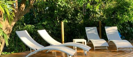 A wooden deck with four white lounge chairs and a small table, surrounded by lush greenery and trees.