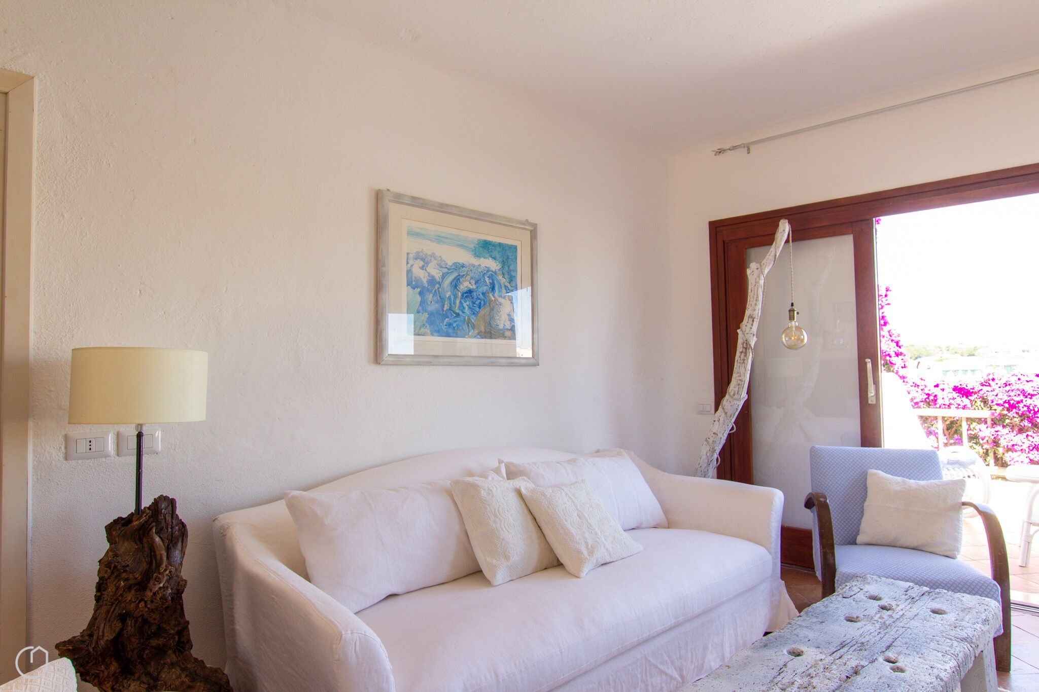 Un sofá blanco con almohadones, una lámpara, un cuadro enmarcado y una silla cerca de una ventana con vistas a flores.