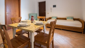 Un coin repas avec une table dressée pour deux personnes, un canapé en osier agrémenté de coussins et une porte visible au fond.