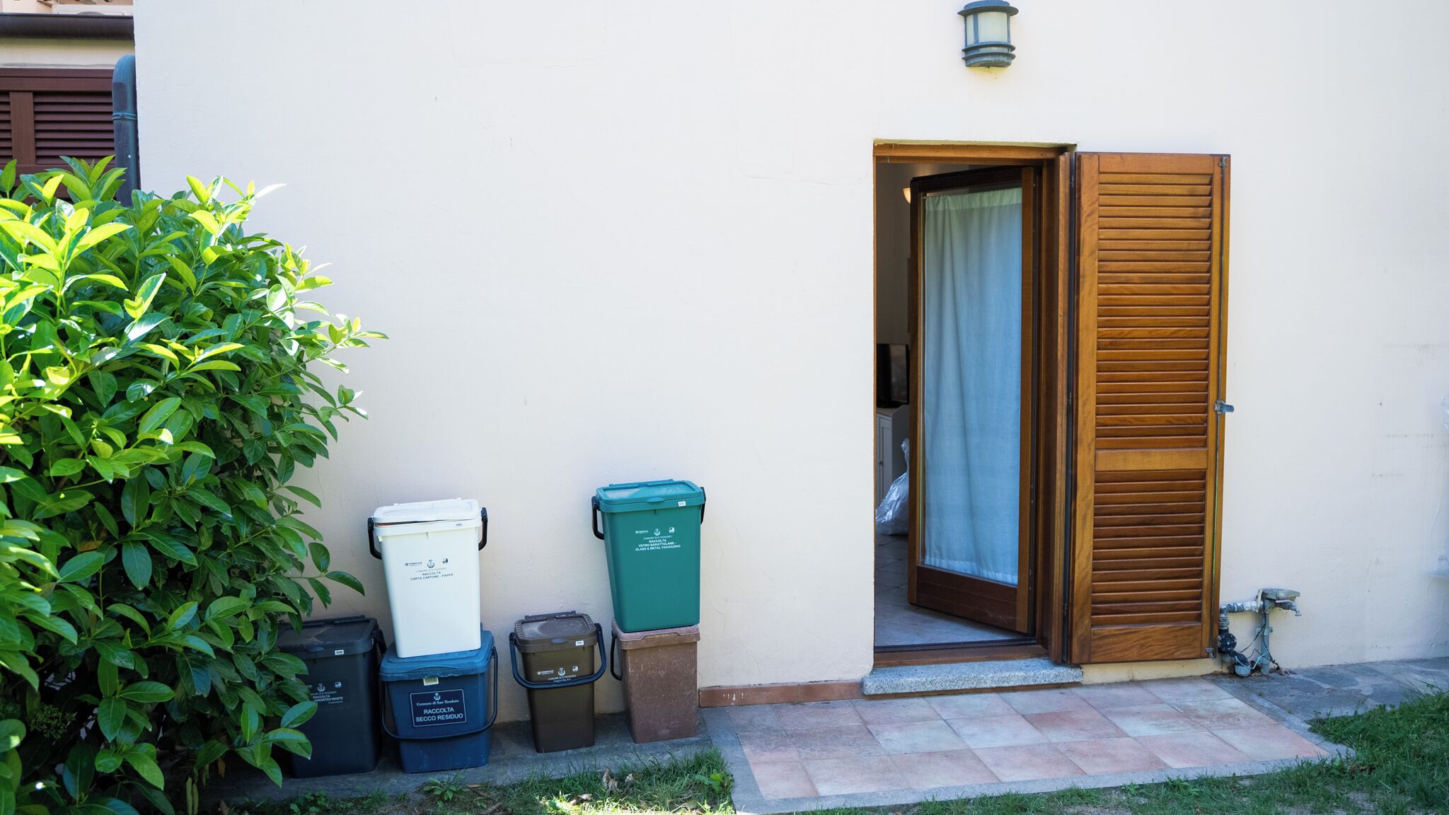 Un'area esterna pulita e ben tenuta, con un muro bianco, una porta in legno e un piccolo giardino con arbusti verdi. Si notano tre cestini per i rifiuti di colori e dimensioni diverse.