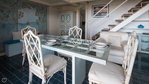 Uma sala de jantar com mesa de vidro, cadeiras brancas e uma escada ao fundo.
