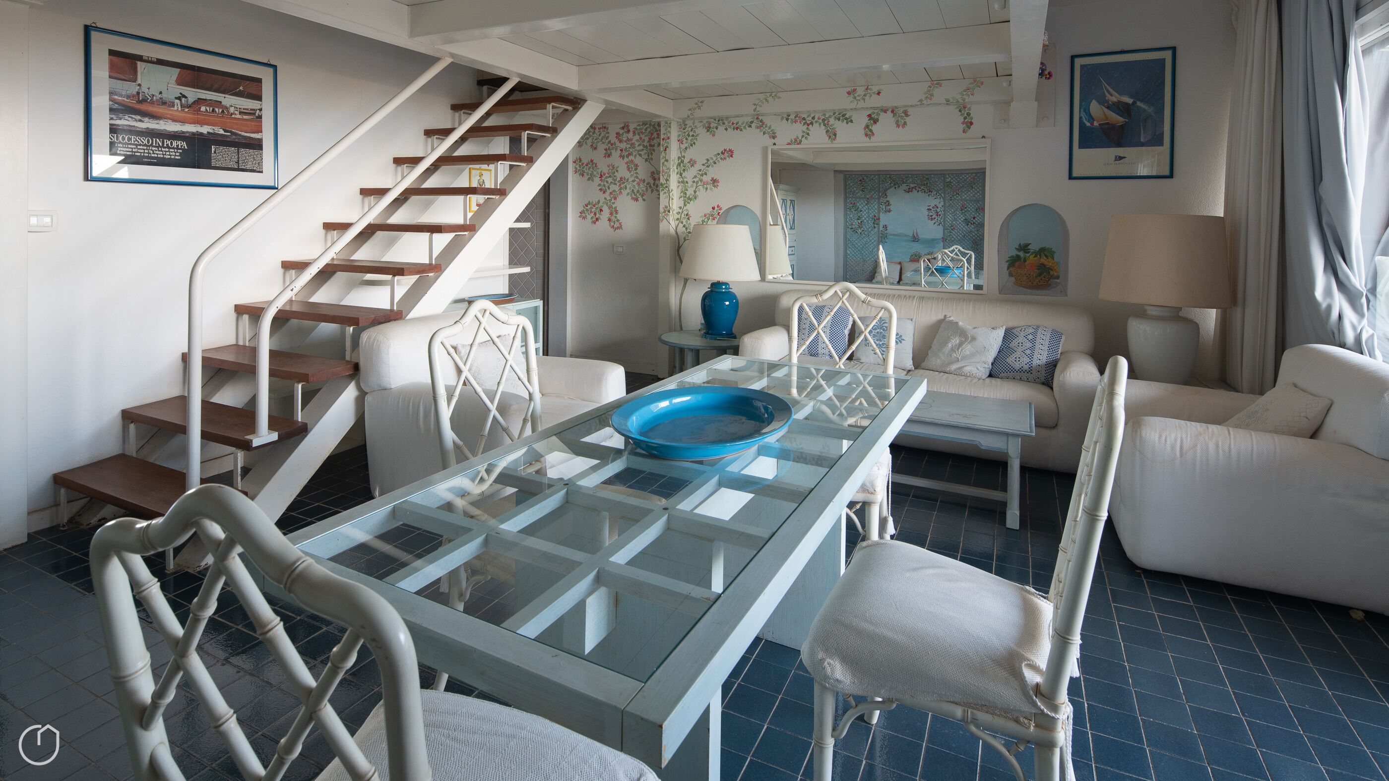 Ein modernes Wohnzimmer mit einem Esstisch mit Glasplatte, weißen Möbeln, einer Treppe und maritimer Dekoration.