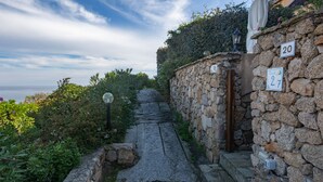 Uma trilha de pedra serpenteia a encosta, ladeada por muros de pedra e vegetação.