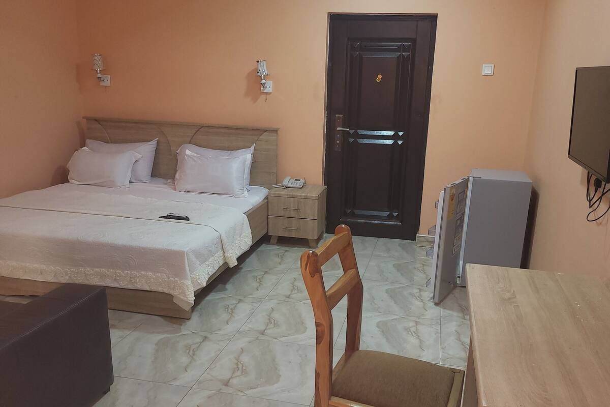 A hotel room with a bed, bedside table, chair, desk, TV, and a door.