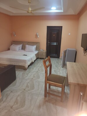 A hotel room with a bed, bedside table, chair, desk, TV, and a door.