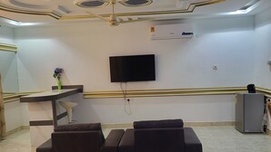 Un salon moderne avec un sol en marbre, un grand téléviseur à écran plat fixé au mur, un ensemble de canapés et un petit réfrigérateur.
