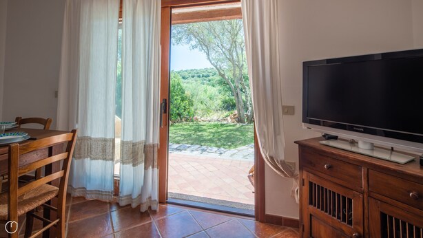 A room with a wooden dining table, chairs, a television on a wooden cabinet, and a sliding glass door leading to a garden. - Mediterranea - One Bedroom Apartment, Sleeps 4 (Loiri)