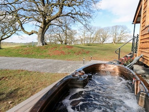 Ein Whirlpool mit einer Holzdar Terrasse, umgeben von einer Rasenfläche mit Bäumen und einem Holzgebäude im Hintergrund.
