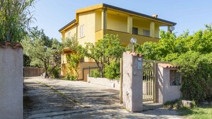 Un bâtiment jaune avec une entrée fermée par un portail, entouré de verdure et d’arbres.