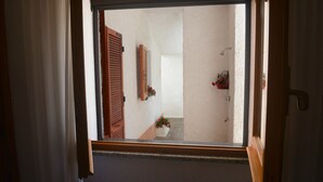 A view through a window into a hallway with a wooden door, a wall-mounted shelf with flowers, and a shower area. - La Casa Di Rita - Salamaghe (Muriscuvò)