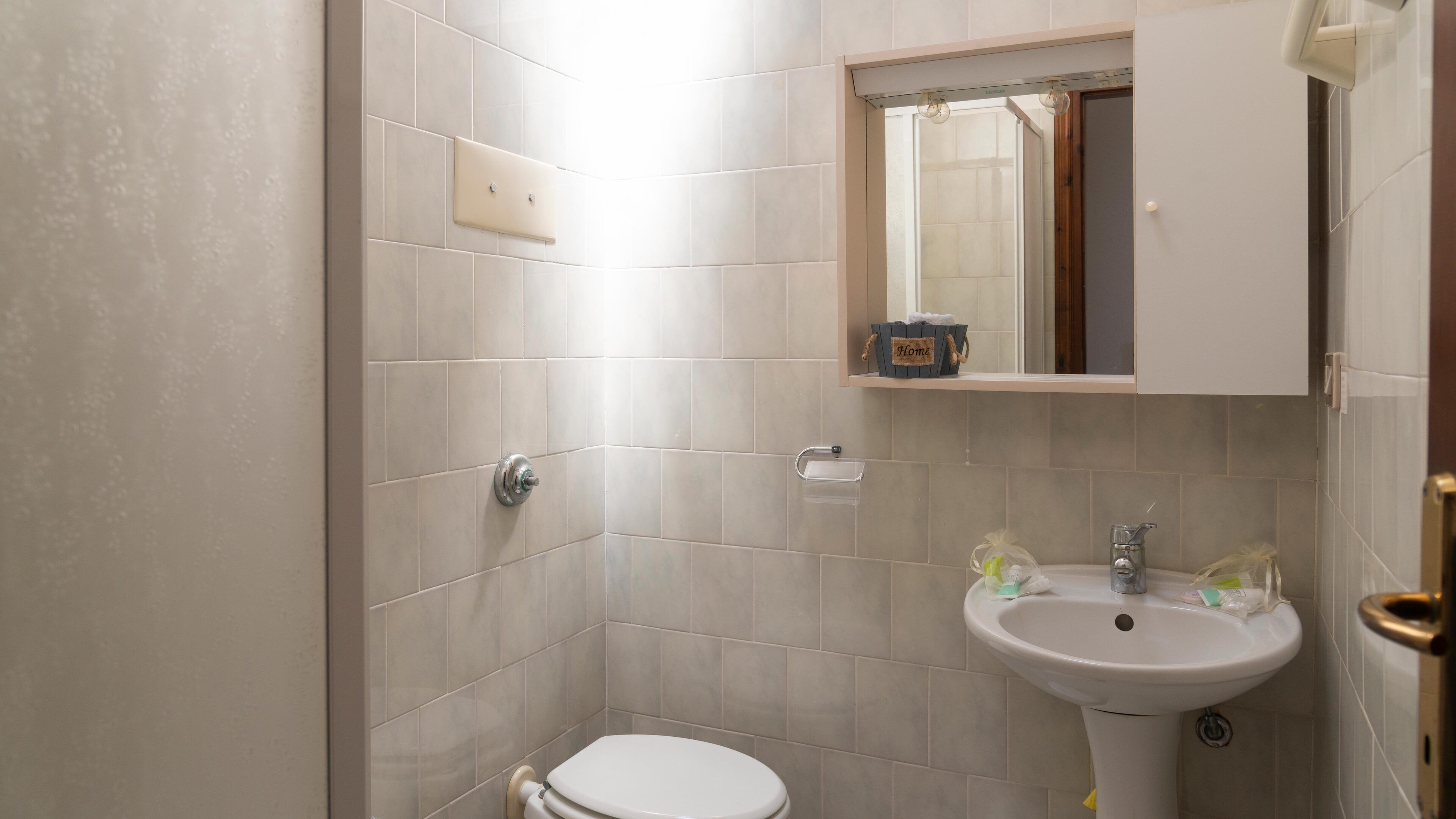 A small bathroom with a toilet, sink, and a mirror above the sink. There is a shelf with a 'Home' sign and a small box.
