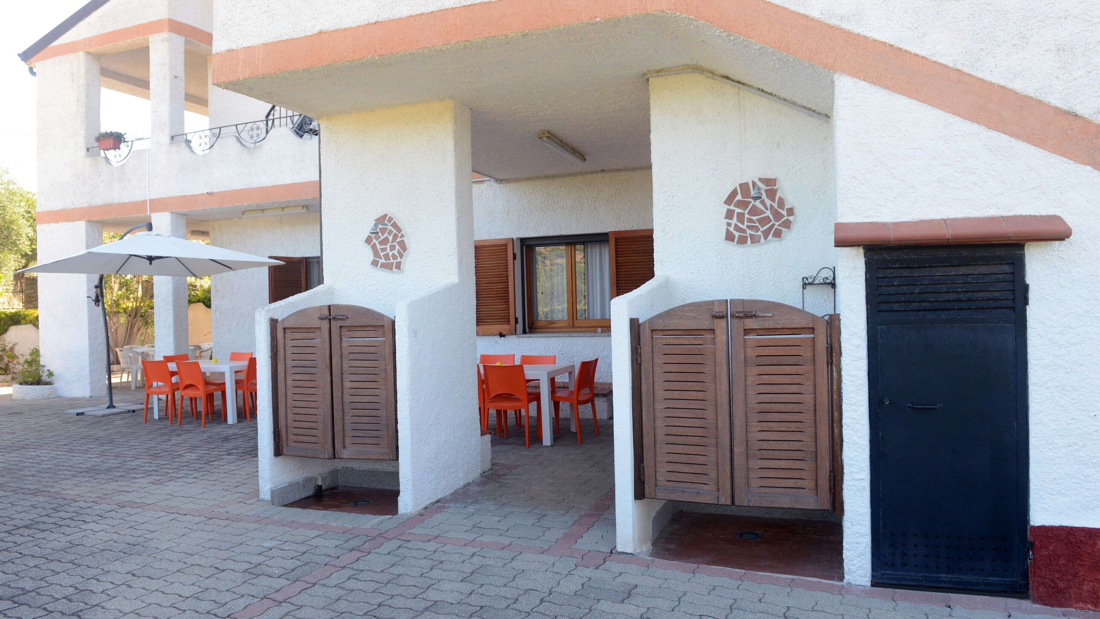 An outdoor patio with tables and chairs, a covered area with an umbrella, and a building entrance with a door and shutters.