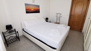 A neatly made bed with white linens, a bedside table with a lamp, and a wall-mounted picture of a sunset. - La Casa Di Rita - A - Sa Capannizza (Muriscuvò)