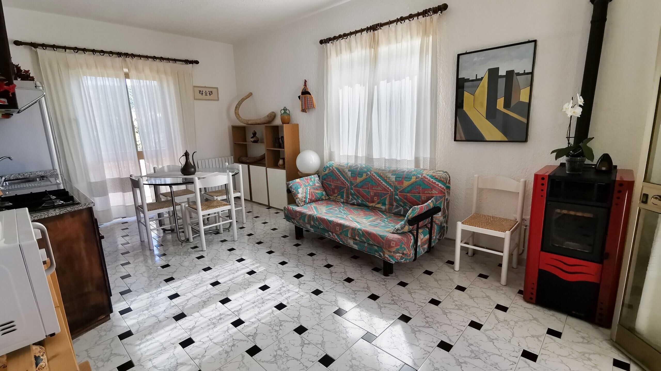 A living room with a patterned sofa, a dining table with chairs, a kitchen area with a stove, and a red appliance.