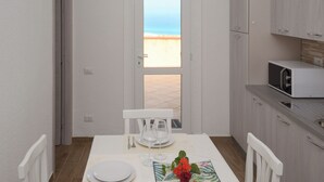 A dining area with a table set for two, a view of the beach through an open door, and a kitchenette with a microwave. - Villa Paola - Panoramic Seaview Terrace (Costa Rei)
