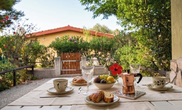 Mesa al aire libre con diversos alimentos y bebidas, una cafetera y vista a un edificio al fondo.