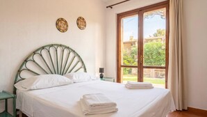 A bedroom with a bed, a nightstand, a window with a view of greenery, and two wall decorations. - Villa Giorgia with garden - 50m from the Beach (Costa Rei)