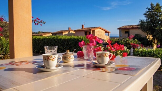 A table with a teapot, cups, and saucers, set outdoors with a view of buildings and greenery. - Villa Oleandro with garden & BBQ - near the beach (Costa Rei)