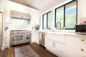 A modern kitchen with white cabinetry, stainless steel appliances, and a large window offering a view of a forested area. - Relaxing country getaway in your dream house (Pemberton)