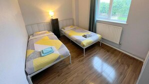 Ett litet, väl upplyst rum med två enkelsängar, ett element och ett fönster med gardiner.