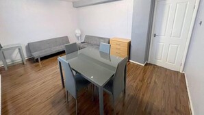 Ett litet, modernt rum med ett matbord med glasskiva, grå stolar, en grå soffa, ett träskåp och en vit dörr.