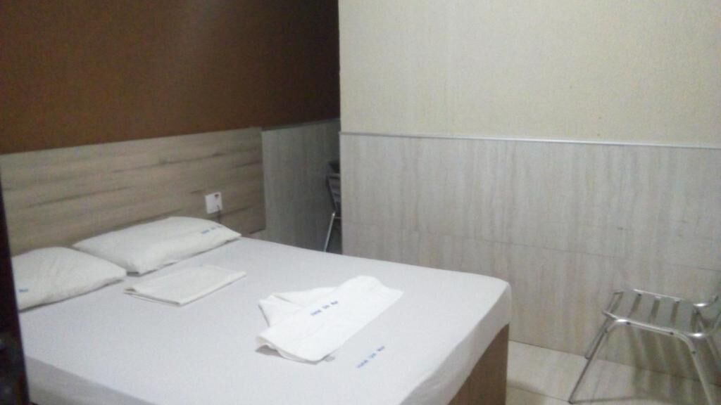 Quarto de hotel com cama, uma cadeira e um porta-toalhas fixado na parede.