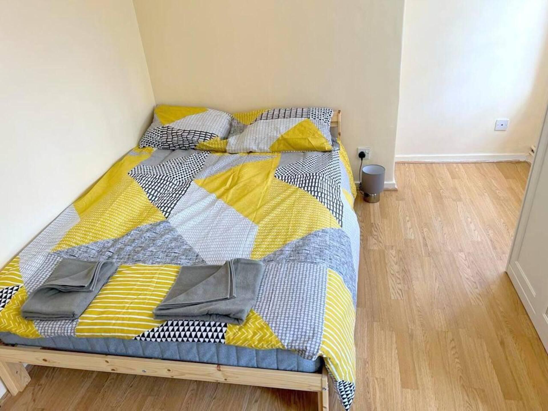 Un lit simple recouvert d’une couvre-lit aux motifs géométriques dans les tons de jaune, de gris et de blanc, agrémenté d’oreillers aux motifs similaires. La pièce est dotée d’un plancher en bois et d’un mur uni avec une prise de courant.