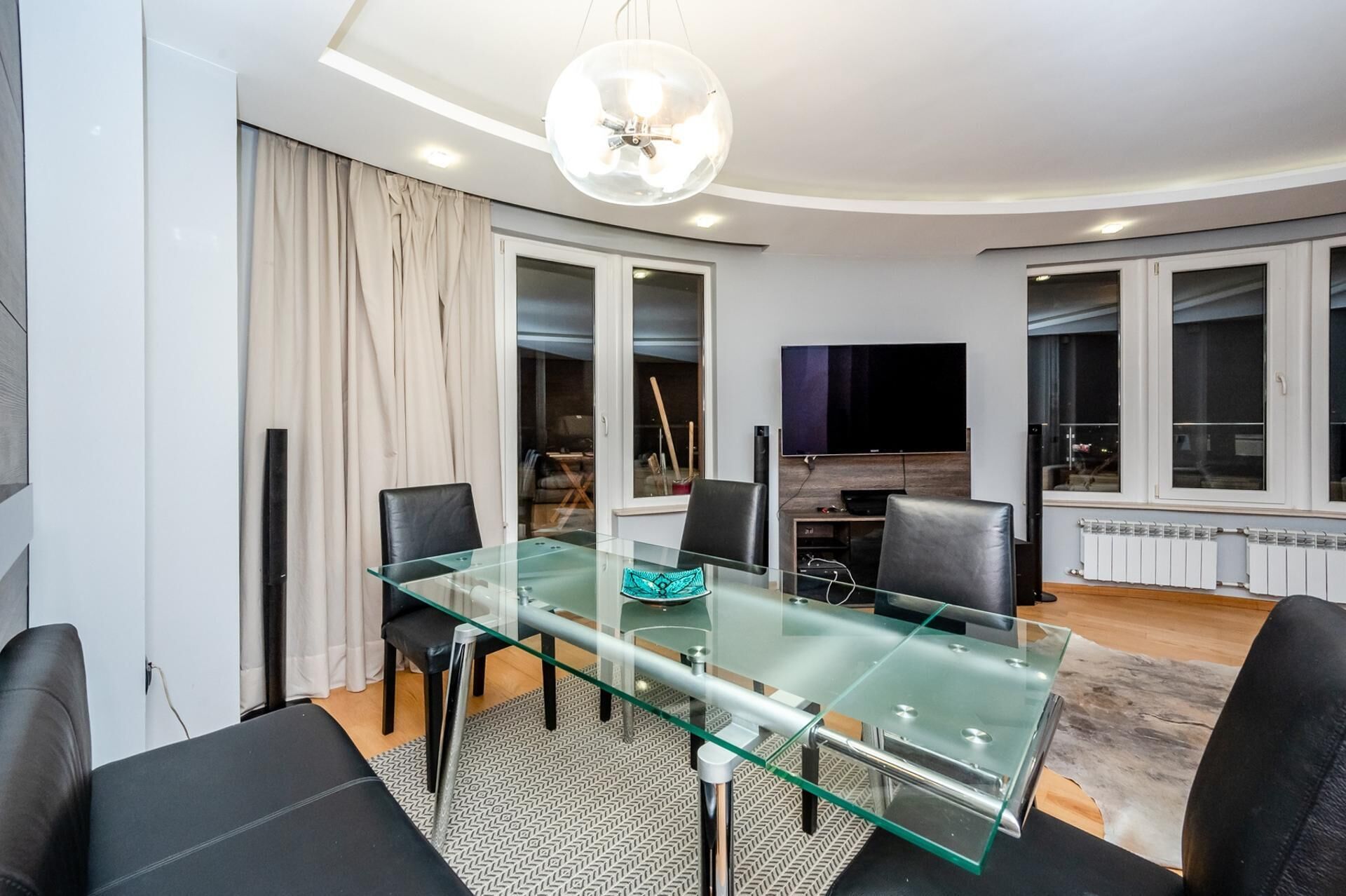 Un salon moderne avec une table à manger en verre, des fauteuils en cuir noir, un grand téléviseur à écran plat et un luminaire suspendu au plafond.
