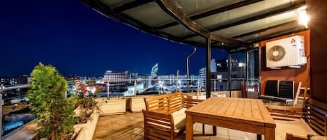 Une terrasse extérieure avec une table et des chaises, un toit en verre et une vue sur les toits de la ville illuminés la nuit.