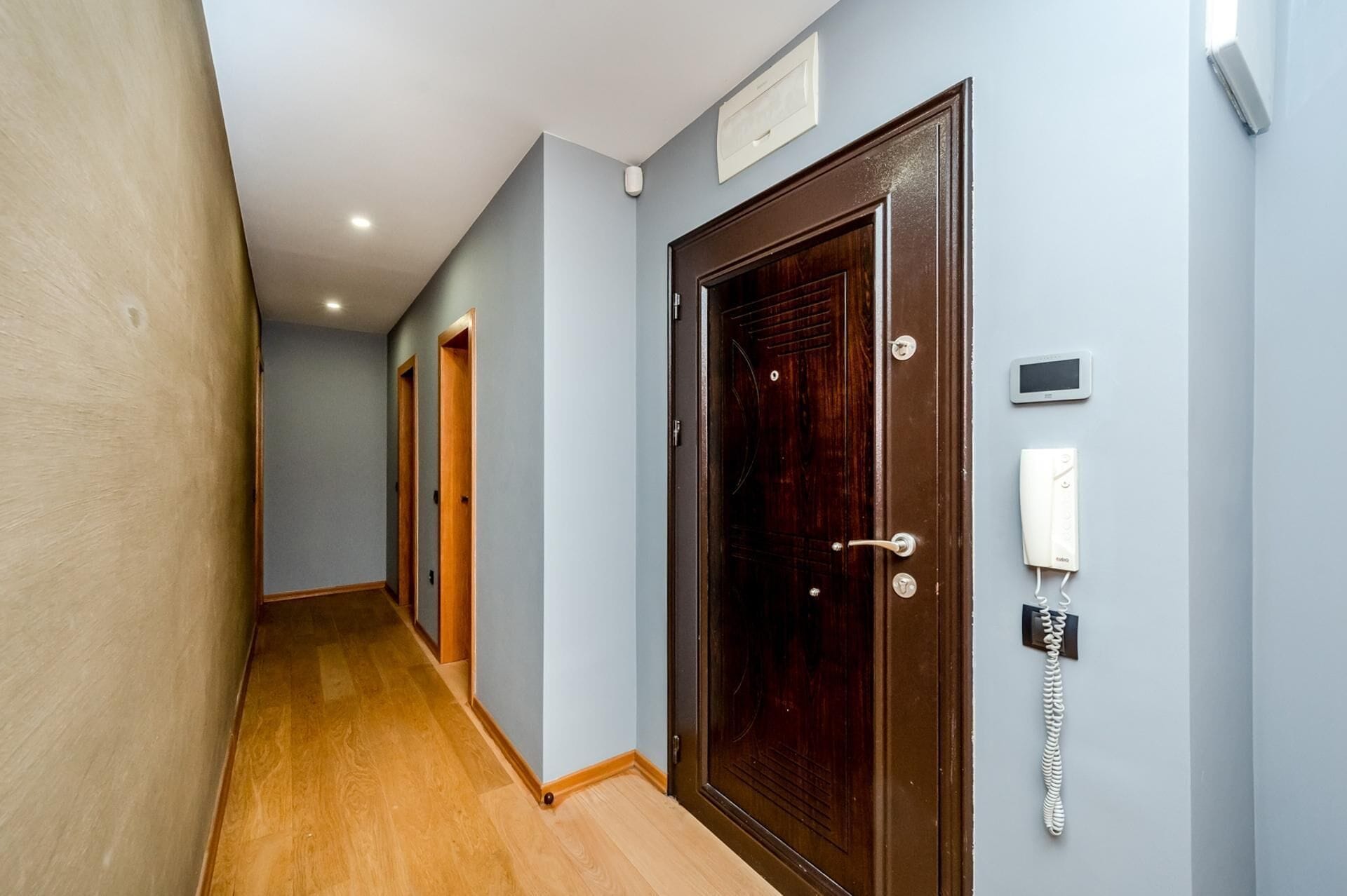 A hallway with wooden flooring, a dark wooden door, a wall-mounted telephone, and a light switch.